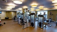 This fitness center is thoughtfully designed to offer a complete workout experience within a single, open-plan floor. The space is outfitted with a full suite of Precor brand equipment, including multiple cable and strength machines arranged along the central floor, as well as cardio machines such as treadmills and ellipticals positioned along the windowed perimeter. Large windows along one wall allow natural light to fill the room, creating a bright and energizing atmosphere. Overhead, a series of distinctive round drum-style pendant lights are evenly distributed across the ceiling, providing consistent, warm illumination throughout the space. The ceiling itself features a subtle dark finish, which adds depth and contrast to the overall design. Walls are painted in a muted olive-green tone, contributing to a calm and focused environment. The flooring is a durable, neutral-toned material suited for high-traffic fitness use. The layout is open and well-organized, allowing users to move freely between equipment. The overall ambiance is polished and professional, reflecting a facility designed with both functionality and aesthetics in mind.