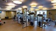This fitness center is thoughtfully designed to offer a complete workout experience within a single, open-plan floor. The space is outfitted with a full suite of Precor brand equipment, including multiple cable and strength machines arranged along the central floor, as well as cardio machines such as treadmills and ellipticals positioned along the windowed perimeter. Large windows along one wall allow natural light to fill the room, creating a bright and energizing atmosphere. Overhead, a series of distinctive round drum-style pendant lights are evenly distributed across the ceiling, providing consistent, warm illumination throughout the space. The ceiling itself features a subtle dark finish, which adds depth and contrast to the overall design. Walls are painted in a muted olive-green tone, contributing to a calm and focused environment. The flooring is a durable, neutral-toned material suited for high-traffic fitness use. The layout is open and well-organized, allowing users to move freely between equipment. The overall ambiance is polished and professional, reflecting a facility designed with both functionality and aesthetics in mind.