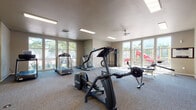 This fitness center at 4823–4841 Aster St in Springfield, Oregon, offers a spacious, single-floor layout designed to accommodate a wide range of workout routines. The room is finished with neutral taupe walls and durable low-pile carpet flooring, creating a practical and comfortable exercise environment. Recessed rectangular fluorescent light panels are evenly distributed across the ceiling, supplemented by a ceiling fan that promotes air circulation throughout the space.

Natural light is a standout feature, with large multi-pane windows lining multiple walls and providing views of the surrounding outdoor areas, including a nearby playground visible from certain vantage points. This abundance of natural light gives the room an open and inviting atmosphere.

The fitness center is well-stocked with a diverse array of equipment, including multiple treadmills, elliptical trainers, a rowing machine, a stationary recumbent bike, a cable weight machine, free weight benches, and a stability ball. A snack and beverage vending machine is conveniently positioned in the corner. Informational notices and fitness guidelines are posted on the walls. Glass-paneled doors connect to an adjacent room, further enhancing the sense of openness within the facility.