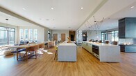 Located at 1835 Morehead Ridge Dr in Charlotte, NC, this single-floor amenity space delivers a polished, contemporary design tailored for communal living and gathering. The open-plan layout seamlessly connects a fully equipped kitchen, dining area, and multiple lounge zones, all unified by warm hardwood-style flooring and soft, neutral wall tones throughout.

The kitchen is a standout feature, showcasing twin quartz-topped islands with ample seating, sage-green cabinetry with dark hardware, built-in ovens, beverage refrigerators, and an artistic multi-pendant light fixture that serves as a sculptural focal point. Adjacent to the kitchen, a generous dining area accommodates a large communal table surrounded by mixed modern seating.

The lounge areas are thoughtfully arranged to suit different social needs. A dramatic three-sided glass fireplace anchors the central living space, creating an inviting atmosphere visible from multiple vantage points. Floor-to-ceiling black-framed windows flood the seating areas with natural light while offering views of the surrounding community. A more intimate lounge nook features plush armchairs, a marble-topped coffee table, and a wall-mounted television. The entry area includes elevator access and direct outdoor connectivity, enhancing the flow of the space. Overall, the design balances modern sophistication with comfortable functionality.
