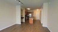 This single-floor space offers a well-designed open-concept layout that connects the kitchen, dining, and living areas into one cohesive and functional environment. Rich hardwood flooring runs throughout, providing warmth and continuity across the space.

The kitchen is a standout feature, showcasing medium-toned wood cabinetry paired with dark granite countertops and a full suite of stainless steel appliances, including a refrigerator and range. A central kitchen island adds additional counter space and serves as a natural gathering point. Track lighting above the kitchen enhances both task and ambient illumination.

Adjacent to the kitchen, a built-in shelving nook and a breakfast bar or pass-through counter offer added storage and flexibility. Flush-mount ceiling fixtures provide consistent lighting throughout the common areas.

The open living area is generously proportioned, benefiting from a clean, unobstructed floor plan that allows for versatile furniture arrangements. Neutral wall tones in warm beige complement the overall palette and create a bright, inviting ambiance. A corridor leading toward the entry door suggests additional private rooms beyond the main living space. The front entry is well-defined, with a solid door and access panel nearby, offering a clear and organized transition into the unit.