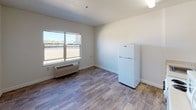 This single-floor studio offers a practical and well-organized layout designed to maximize every square foot. The main living area features durable wood-look flooring throughout and is bathed in natural light from a large window that overlooks the surrounding exterior. A wall-mounted HVAC unit ensures year-round climate comfort, and a full-size refrigerator provides ample food storage.

The kitchen is efficiently arranged along one wall and includes a four-burner electric range, a single-basin sink with cabinetry below, a microwave, and additional counter space. White cabinetry keeps the aesthetic clean and cohesive, complementing the neutral tones found throughout the unit.

A dedicated alcove area offers a built-in desk surface, ideal for a workspace or study area, alongside a tall two-door storage cabinet that provides excellent organizational capacity. Adjacent to this space is a walk-in closet area, adding meaningful storage for clothing and personal belongings.

The bathroom features a full-size soaking tub and shower combination with chrome fixtures and a clean, all-white surround, maintaining the unit's consistent neutral palette. Textured ceilings add a subtle layer of architectural detail throughout the space. Overall, the unit presents a functional, move-in-ready environment with a clean, minimalist character.