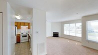 This one-bedroom space is laid out across a single floor, offering a functional and comfortable design. The living area is finished with plush wall-to-wall carpeting and is anchored by a fireplace, providing a warm focal point for the room. A large window allows natural light to fill the space, creating a bright and welcoming atmosphere with views of the surrounding outdoor scenery.

The kitchen is positioned adjacent to the living area, separated by a partial wall that helps define each space while maintaining an open feel. It features tile flooring, classic oak cabinetry, and white appliances including an electric range, offering a practical and tidy cooking environment. A range hood is mounted above the stovetop, adding to the kitchen's functionality.

Throughout the space, the walls are finished in a clean, neutral white tone that complements both the warm wood cabinetry and the light carpeting in the living area. A thermostat and floor-level ventilation register are also visible, indicating a central climate control system. Overall, the space offers a clean, move-in ready layout suited for comfortable everyday living.