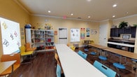 This inviting single-floor educational space is thoughtfully designed to foster learning and creativity in a child-friendly environment. The main room is finished with warm yellow walls and rich dark hardwood flooring, creating a cheerful yet grounded atmosphere filled with natural and recessed lighting.

The space is organized to support various learning activities. Long white tables paired with colorful chairs in shades of yellow, orange, and teal provide ample room for group work and collaborative projects. A separate seating area with round tables and stools offers additional space for smaller group activities or individual study.

A large, well-stocked bookshelf lines one wall, filled with an extensive collection of children's books and educational materials, making it a central resource hub for young learners. Bright educational posters and colorful wall decorations — including color charts, butterfly motifs, and themed displays — adorn the walls, adding visual stimulation and reinforcing key learning concepts.

Crown molding frames the ceiling, lending a polished, finished look to the room. Multiple doors provide easy access and flow throughout the space. Overall, the layout is open, organized, and purposefully designed to create an engaging, energetic, and welcoming environment for children's education and enrichment activities.