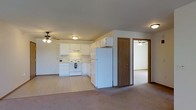 This single-floor space offers an open-concept layout that seamlessly connects the kitchen and living areas. The kitchen is outfitted with white cabinetry providing ample storage, a white electric range, and a full-size refrigerator. A range hood is positioned above the stove, and the countertops offer functional prep space. The kitchen flooring features light-toned tile, which transitions into wall-to-wall carpeting in the adjacent living area, creating a clear visual distinction between the two zones.

A ceiling fan is installed in the living area, promoting air circulation throughout the space. The kitchen area also features a ceiling-mounted light fixture for consistent illumination. Neutral wall tones contribute to a clean, understated aesthetic throughout.

A hallway visible from the living area leads to additional rooms, including what appears to be a bedroom accessible through a wood-toned door. A second ceiling fan is visible in that adjoining room, suggesting comfortable climate control throughout. The layout offers a straightforward and practical flow between living, kitchen, and private spaces.