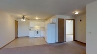This single-floor space offers an open-concept layout that seamlessly connects the kitchen and living areas. The kitchen is outfitted with white cabinetry providing ample storage, a white electric range, and a full-size refrigerator. A range hood is positioned above the stove, and the countertops offer functional prep space. The kitchen flooring features light-toned tile, which transitions into wall-to-wall carpeting in the adjacent living area, creating a clear visual distinction between the two zones.

A ceiling fan is installed in the living area, promoting air circulation throughout the space. The kitchen area also features a ceiling-mounted light fixture for consistent illumination. Neutral wall tones contribute to a clean, understated aesthetic throughout.

A hallway visible from the living area leads to additional rooms, including what appears to be a bedroom accessible through a wood-toned door. A second ceiling fan is visible in that adjoining room, suggesting comfortable climate control throughout. The layout offers a straightforward and practical flow between living, kitchen, and private spaces.