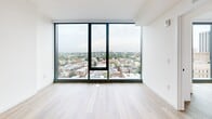 Located at 801 N Orange St in the heart of Wilmington, Delaware, this contemporary high-rise unit offers a thoughtfully designed open-concept layout across a single floor. The combined kitchen and living area serves as the central hub of the space, featuring warm light wood flooring that flows throughout. The modern kitchen is outfitted with sleek gray cabinetry extending to the ceiling for ample storage, white quartz countertops, a large central island with pendant lighting, and a full suite of stainless steel appliances including a refrigerator, range, microwave, and dishwasher. Floor-to-ceiling windows in the living area flood the space with natural light and frame breathtaking panoramic views of the Wilmington cityscape, including historic architecture and the broader urban landscape. The bathroom is elegantly finished with large-format tile flooring, a soaking tub-shower combination clad in dramatic marble-style tile, and a wood-accented vanity with a white countertop. The generously sized walk-in closet provides extensive built-in shelving and double hanging rods, offering exceptional organizational flexibility. Clean white walls and a minimalist aesthetic create a bright, modern ambiance throughout.