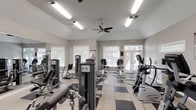 This fitness center offers a comprehensive workout environment designed to accommodate a variety of exercise routines. The space features vaulted ceilings that contribute to an open, airy atmosphere, while ceiling fans ensure comfortable air circulation throughout. The flooring consists of a distinctive two-tone tile pattern in gray and white, adding a clean, modern aesthetic to the room.

Large windows fitted with white blinds line the walls, allowing abundant natural light to brighten the interior and providing views of the surrounding greenery. A set of glass double doors at the far end further enhances the connection between the indoor space and the outdoors.

The fitness center is outfitted with an impressive array of equipment. The strength training section features multiple cable and resistance machines positioned along a mirrored wall, allowing users to monitor their form during workouts. The cardio section includes stationary bikes, elliptical trainers, and additional aerobic equipment arranged to maximize floor space and ease of movement.

Overall, the space strikes a balance between functionality and comfort, offering a bright, well-organized environment suitable for both casual and dedicated fitness routines. The layout promotes an efficient workflow, ensuring users can transition seamlessly between strength and cardio training.