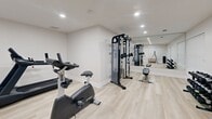 Located at 125 Parker Pl in LaGrange, Georgia, this dedicated exercise room offers a clean, purposefully designed fitness space. The room features light-toned hardwood-style flooring that brightens the interior and complements the crisp white walls and white baseboards, creating a fresh and motivating atmosphere.

Recessed ceiling lighting provides consistent, even illumination throughout the space, while a large full-wall mirror along one side visually expands the room and supports form-checking during workouts. The mirrored wall reflects the full array of equipment, enhancing the sense of openness. A cable machine, stationary bike, elliptical, weight bench, and dumbbell rack are among the fitness equipment present, offering a well-rounded range of cardio and strength training options.

A glazed entry door with a welcome mat and sidelight windows allows natural light to filter in from outside, adding a welcoming touch to the functional environment. Horizontal blinds on the windows offer privacy and light control as needed. Safety features include a mounted fire extinguisher and an illuminated exit sign near the entrance.

A wooden bench provides a convenient spot for stretching or resting between sets, and a sleek waste bin adds to the room's practical amenities. This exercise room is efficiently arranged to maximize usability within its compact footprint.