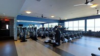 Located at 145 Westerly Rd in Ossining, NY, this fitness center offers a thoughtfully designed, single-floor layout built to support a comprehensive workout experience. The cardio floor is expansive, lined with rows of modern equipment including treadmills, ellipticals, and stationary bikes, all arranged to maximize space and usability. Bold blue accent walls contrast with light wood-toned flooring, creating an energetic yet clean aesthetic throughout the space. Large floor-to-ceiling windows along one wall flood the interior with natural light while offering sweeping views of the surrounding waterfront landscape, providing a compelling backdrop during workouts. Ceiling-mounted fans are evenly distributed across the room, ensuring air circulation and comfort during high-intensity exercise sessions. Full-length mirrors along the walls enhance the sense of openness and allow users to monitor their form. Recessed and flush-mount lighting supplements the natural light, keeping the space well-illuminated at all hours. The overall ambiance is modern and motivating, combining functional design with an inviting atmosphere suited for fitness enthusiasts of all levels.