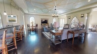 This sophisticated single-floor space showcases a refined blend of classical and contemporary design elements throughout. The expansive main living area commands immediate attention with its striking coffered ceiling, painted in soft blue and accented with ornate gold-toned molding, anchored by a wrought iron chandelier that casts warm, ambient light across the room. Rich, dark hardwood flooring flows continuously throughout, grounding the space with a sense of warmth and elegance.

Dramatic arched windows bathe the interiors in generous natural light while offering views to the outdoors, complemented by a stunning Gothic-arched wrought iron decorative gate that serves as a bold architectural focal point. Decorative columns further define the open layout, subtly separating the seating areas without sacrificing the spacious, airy feel.

The living area is thoughtfully arranged with multiple comfortable seating groupings, accommodating both intimate conversation and larger gatherings. An integrated dining nook, framed by arched windows, provides a bright and inviting space for meals.

The kitchen features a generous island with dark countertops and raised bar seating, cream-toned cabinetry, and pendant lighting that highlights the functional workspace. An arched window above the counter area ensures this room also benefits from ample natural light, creating a seamless connection to the overall warmth and character of the space.