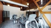 This distinctive single-floor facility masterfully blends historic architectural character with modern functionality. The space is housed within what appears to be a converted barn or heritage building, highlighted by dramatic exposed wood-beam ceilings, warm tongue-and-groove wood paneling overhead, and soaring interior heights that create an open, airy atmosphere throughout.

The fitness area is a standout feature, equipped with a comprehensive selection of cardio machines including treadmills, ellipticals, and stationary bikes, as well as medicine balls for functional training. Durable carpet tiles cover the exercise floor, while exposed ductwork and pendant globe lighting add an industrial-chic aesthetic. A mezzanine-level balcony with dark-stained wooden railings overlooks the fitness space below, reinforcing the building's striking vertical scale.

A lounge and entry area provides a welcoming transition into the space, featuring warm hardwood-style flooring, a dark-stained staircase leading to the mezzanine, decorative wall accents, a bookshelf, and water fountains. The lounge is furnished with navy blue upholstered seating, colorful accent pillows, and a vibrant teal-toned botanical canvas artwork that adds a pop of color. A kitchenette area with dark cabinetry and a light countertop completes the social space, making this facility both functional and inviting.