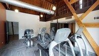 This distinctive single-floor facility masterfully blends historic architectural character with modern functionality. The space is housed within what appears to be a converted barn or heritage building, highlighted by dramatic exposed wood-beam ceilings, warm tongue-and-groove wood paneling overhead, and soaring interior heights that create an open, airy atmosphere throughout.

The fitness area is a standout feature, equipped with a comprehensive selection of cardio machines including treadmills, ellipticals, and stationary bikes, as well as medicine balls for functional training. Durable carpet tiles cover the exercise floor, while exposed ductwork and pendant globe lighting add an industrial-chic aesthetic. A mezzanine-level balcony with dark-stained wooden railings overlooks the fitness space below, reinforcing the building's striking vertical scale.

A lounge and entry area provides a welcoming transition into the space, featuring warm hardwood-style flooring, a dark-stained staircase leading to the mezzanine, decorative wall accents, a bookshelf, and water fountains. The lounge is furnished with navy blue upholstered seating, colorful accent pillows, and a vibrant teal-toned botanical canvas artwork that adds a pop of color. A kitchenette area with dark cabinetry and a light countertop completes the social space, making this facility both functional and inviting.