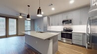 This thoughtfully designed single-floor residence offers a clean, contemporary aesthetic throughout. The heart of the home is an open-concept kitchen that commands attention with its large center island topped in light quartz, equipped with an undermount sink and accented by stylish pendant lighting. Gray shaker-style cabinetry lines the walls from floor to ceiling, complemented by crisp white countertops and a full suite of stainless steel appliances, including an electric range and over-the-range microwave. Wood-look plank flooring flows seamlessly through the kitchen and living areas, adding warmth to the modern palette. The bedroom is bright and welcoming, featuring dual windows with horizontal blinds that invite natural light into the neutral-toned space. The primary bathroom is generously proportioned, showcasing large-format light tile flooring, a warm wood-toned vanity with a white countertop, a well-lit mirror, and direct access to a spacious walk-in closet with built-in shelving. A glass-enclosed shower adds a refined touch. The layout, as revealed in the dollhouse view, is efficiently organized, connecting the open living and kitchen area to the private bedroom and bathroom wing through a functional corridor. Overall, the space presents a clean, move-in-ready environment with modern finishes and practical design.