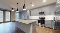 This thoughtfully designed single-floor residence offers a clean, contemporary aesthetic throughout. The heart of the home is an open-concept kitchen that commands attention with its large center island topped in light quartz, equipped with an undermount sink and accented by stylish pendant lighting. Gray shaker-style cabinetry lines the walls from floor to ceiling, complemented by crisp white countertops and a full suite of stainless steel appliances, including an electric range and over-the-range microwave. Wood-look plank flooring flows seamlessly through the kitchen and living areas, adding warmth to the modern palette. The bedroom is bright and welcoming, featuring dual windows with horizontal blinds that invite natural light into the neutral-toned space. The primary bathroom is generously proportioned, showcasing large-format light tile flooring, a warm wood-toned vanity with a white countertop, a well-lit mirror, and direct access to a spacious walk-in closet with built-in shelving. A glass-enclosed shower adds a refined touch. The layout, as revealed in the dollhouse view, is efficiently organized, connecting the open living and kitchen area to the private bedroom and bathroom wing through a functional corridor. Overall, the space presents a clean, move-in-ready environment with modern finishes and practical design.