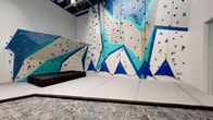Located on S Alton St in Denver, Colorado, this single-floor fitness facility offers a compelling combination of athletic amenities and thoughtfully designed communal spaces. The standout feature is the expansive indoor rock climbing area, where soaring walls are adorned with colorful geometric patterns in bold shades of teal, blue, and white. Multiple routes are set across angled and vertical surfaces, accommodating a range of skill levels. Thick safety padding lines the base of the climbing walls, and patterned carpet flooring defines the surrounding spectator and rest areas, where modular seating provides a comfortable place to observe or recover.

Adjacent to the climbing zone, a fully equipped fitness room is visible through sleek floor-to-ceiling glass partitions, housing a variety of strength and resistance machines beneath bright, even lighting. This transparent boundary keeps the space feeling open and connected while clearly delineating each functional zone.

A separate lounge area features light hardwood flooring, soft organic-shaped seating, and glass-walled rooms that reveal an elegant interior staircase with wood-paneled walls. Recessed lighting throughout lends a clean, polished aesthetic. The overall design balances high-energy athletic functionality with a modern, welcoming ambiance.