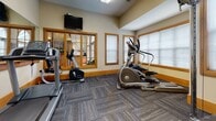 Located at 7260 Cold Harbor Rd in Mechanicsville, Virginia, this fitness center offers a well-equipped, single-floor workout facility designed to support a comprehensive exercise routine. The space is thoughtfully laid out to accommodate both cardiovascular and strength training needs.

The cardio area features a treadmill, elliptical trainer, and Matrix recumbent stationary bike, providing a variety of low- and high-impact workout options. A multi-station cable machine serves as the centerpiece of the strength training area, complemented by a workout reference chart posted on the wall for exercise guidance.

The room is finished with durable, patterned carpet tiles in a neutral gray tone, warm wood trim accents, and cream-colored walls that create a clean, welcoming atmosphere. Large windows fitted with blinds allow natural light to fill the space, enhancing the open feel of the room. A full-length wall mirror visually expands the area and allows users to monitor their form during workouts. A wall-mounted flat-screen television adds an entertainment option for those using the cardio equipment.

Interior windows connect the fitness room to an adjacent space, adding architectural interest and a sense of openness. Overhead fluorescent lighting ensures the room remains well-lit throughout the day. Sanitation wipes are conveniently available, reflecting a commitment to cleanliness and user comfort.