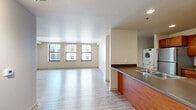 This single-floor unit offers a thoughtfully designed open-concept layout that maximizes the generous square footage. The combined kitchen and living area forms the heart of the space, featuring light wood-tone luxury vinyl plank flooring that flows seamlessly throughout. Large windows line the living area, allowing abundant natural light to fill the room while offering views of the surrounding urban streetscape.

The kitchen is well-equipped with warm-toned wood cabinetry, stainless steel appliances including a range and refrigerator, and a granite-style countertop that extends along a peninsula with a built-in sink — ideal for meal prep and casual dining. A stacked washer and dryer unit is conveniently integrated into the kitchen area, adding practical in-unit laundry functionality.

The living area is open and expansive, with clean white walls and a neutral palette that lends a bright, airy atmosphere. The layout flows naturally from the kitchen peninsula through an architectural pass-through, creating defined zones while maintaining an open feel.

The bathroom features tile flooring, a light wood vanity with an integrated sink, a well-lit mirror with overhead lighting, and an adjacent walk-in closet with open wire shelving for ample storage. The overall design is clean, functional, and modern, with a neutral color scheme that adapts easily to a variety of interior styles.