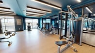 This contemporary fitness center offers a thoughtfully designed, single-floor facility divided into two distinct functional areas. The main workout space is generously proportioned, providing ample room for a comprehensive range of exercise equipment, including cable machines, a power rack with free weights, benches, and cardio equipment such as ellipticals and rowing machines. Large floor-to-ceiling mirrors line the walls, visually expanding the space and allowing users to monitor their form during workouts.

A separate, glass-partitioned studio area serves as a dedicated space for yoga, stretching, and flexibility training. This room features a clean, open layout with hardwood-style flooring, a workout mat, stability balls, and a wall of mirrors, all illuminated by natural light streaming through large windows that overlook the outdoors.

Throughout the facility, the design is unified by bold teal-painted walls, dark coffered ceilings, and rows of recessed linear LED lighting that create a bright, energetic ambiance. A wall-mounted flat-screen television adds a multimedia element to the training environment. The light wood-tone flooring, transitioning to rubberized matting in the strength zone, ensures both aesthetic appeal and functional safety. The overall atmosphere is modern, clean, and purpose-built for focused fitness.