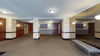 This building lobby presents a well-appointed and professional environment designed to make a strong first impression. The space is defined by cream and beige structural columns accented with dark marble base detailing, which adds a touch of elegance and architectural character. Rich, dark wood-paneled elevator doors and wall features provide a warm contrast against the lighter tones of the walls and columns. A continuous dark marble chair-rail band runs along the walls, reinforcing the cohesive and refined design language throughout the area. The flooring is covered in a neutral, textured carpet that adds comfort underfoot while complementing the overall color palette. Overhead, flush-mounted ceiling light fixtures provide even, functional illumination across the space, with additional wall-mounted sconces contributing to a layered lighting scheme. Decorative cityscape artwork is displayed prominently on the walls, adding visual interest and a contemporary touch to the otherwise classic interior. A sleek upholstered bench is positioned near the entrance, offering a convenient seating option for visitors. The layout is open and unobstructed, providing clear sightlines and easy circulation between the elevator banks and the entry points of the lobby.