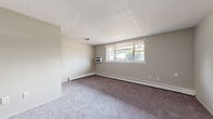 This single-floor residential space offers a practical and functional layout designed for comfortable everyday living. The main living area is generously proportioned, featuring soft wall-to-wall carpeting in a neutral gray tone and light gray painted walls that create a calm, understated atmosphere. A large window along the exterior wall allows natural light to filter in while providing outdoor views of surrounding greenery. The room is equipped with a wall-mounted air conditioning unit and baseboard heating, ensuring year-round climate comfort.

The bedrooms continue the same carpeted flooring and neutral color palette, each offering dedicated closet storage with built-in hanging rods. One bedroom benefits from additional natural light through a window fitted with blinds, while another features convenient access to adjoining spaces.

The kitchen is a compact, efficient galley-style space outfitted with warm-toned wooden cabinetry, a gas range, and a double-basin sink set beneath a window. Granite-patterned countertops provide functional prep space, and durable tile flooring grounds the room.

The bathroom features white full-height wall tiling surrounding a soaking bathtub, complemented by a frosted privacy window that brings in soft diffused light. Overall, the space presents a clean, no-frills interior with practical amenities throughout.