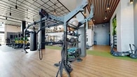 Located at 1600 W Lincoln Ave in Anaheim, California, this two-story fitness facility offers a comprehensive workout environment spread across two generously sized floors. The main level is the larger of the two, characterized by soaring ceilings, a dramatic grid-pattern ceiling design, and floor-to-ceiling glass walls that flood the space with natural light and provide outdoor views of the surrounding landscape. This level houses an extensive functional training zone featuring heavy-duty equipment such as a tire sled, weight racks with colorful bumper plates, cable machines, and turf flooring designated for dynamic exercise. Arched windows add an architectural elegance to the space, complementing the industrial-modern aesthetic created by exposed pendant lighting and bold structural columns. The second floor provides a more intimate exercise environment, equipped with a wide array of cardio machines including stationary bikes, rowing machines, ellipticals, and stair climbers. Full-length mirrors amplify the sense of space, while arched windows overlook the ground level and the exterior. Both floors feature warm wood-look flooring alongside rubber tile zones, ensuring functionality and comfort throughout. A dedicated studio area with a wood-paneled ceiling and glass partition walls adds a refined, multi-purpose dimension to the facility.