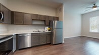 This well-appointed single-floor unit offers a thoughtfully designed open-concept layout that seamlessly connects the kitchen and living areas. The kitchen is a standout feature, showcasing rich dark-toned cabinetry with sleek hardware, light-colored countertops, and a full suite of stainless steel appliances including a refrigerator, range with oven, built-in microwave, and dishwasher. A double-basin sink and tile backsplash add refined, functional detail to the space. Warm-toned wood-look flooring runs throughout the main areas, creating a cohesive and inviting atmosphere. The living area is generously proportioned, benefiting from natural light through a window dressed with horizontal blinds, and features a ceiling fan with light fixture overhead. Recessed lighting in the entry and kitchen areas ensures the space remains bright and well-lit. Multiple interior doors lead to additional rooms and closet storage. The bathroom is clean and modern, featuring a single-sink vanity with matching dark cabinetry, a light stone-style countertop, a large mirror, and a full-size tub-shower combination with shelving. The neutral color palette and consistent finishes throughout give this unit a cohesive, contemporary feel.