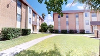 This single-floor unit is part of a multi-building residential complex constructed with classic brick and stucco exteriors. The grounds between the buildings are well-maintained, featuring lush green lawns, manicured shrubs, and mature tropical trees including palms, all connected by a concrete walkway that winds through the property. The overall exterior presents a clean, established appearance set beneath open Florida skies.

Inside, the unit features a consistent and practical design throughout. The interior rooms are finished with light beige textured walls that complement the neutral palette, and light-toned ceramic tile flooring runs throughout the space, providing durability and ease of maintenance. White baseboards offer a clean contrast against the walls and flooring. Each room benefits from windows fitted with horizontal blinds, allowing residents to control the amount of natural light that enters the space. The textured ceilings add dimension to the rooms.

The layout includes multiple rooms separated by doorways framed with white panel doors and detailed door casings, adding a subtle architectural touch to an otherwise straightforward design. The overall ambiance of the interior is clean, neutral, and functional, offering a blank canvas well-suited for personalization.