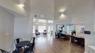 This single-floor community or leasing office is designed to make a strong first impression from the moment you step through the stately wood-and-glass double entry doors. The grand entryway immediately draws the eye upward to dramatically soaring ceilings that lend an airy, expansive feel to the entire space. Transom windows above the entry flood the foyer with natural light, while the warm hardwood-style flooring flows seamlessly throughout.

The open-concept interior is thoughtfully organized into distinct functional zones. A dedicated reception and workstation area, set against a bold teal accent wall and adorned with contemporary artwork, provides a professional yet inviting hub for staff and visitors. Adjacent to this, a welcoming lounge area features plush blue wingback chairs, a glass coffee table, and additional seating arranged to encourage comfortable conversation, all bathed in natural light from large windows with clean white blinds.

A casual dining nook with a round black table and metal chairs offers an informal gathering spot near a secondary glass-paneled door that provides easy access to the exterior. Wall-mounted sconce lighting and recessed fixtures throughout complement the natural light, creating a bright and polished ambiance. The overall design balances a modern, professional aesthetic with warm, approachable furnishings, making this a functional and welcoming space for visitors and staff alike.