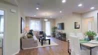 This single-floor residence showcases a welcoming living area defined by warm hardwood flooring that extends throughout the space. The room is illuminated by a combination of recessed ceiling lights and a stylish pendant fixture, ensuring consistent, even lighting throughout. Large windows dressed with sheer and patterned curtains allow generous natural light to filter in, complementing the neutral, warm-toned walls that lend a calm, airy ambiance to the interior.

A glass-paneled door provides access to the outdoors, further enhancing the sense of openness. The living area is thoughtfully arranged with comfortable seating, a dark-framed coffee table set atop a light area rug, and a media console supporting a wall-mounted television. Lush indoor plants are strategically placed throughout, adding a natural, refreshing element to the décor.

The layout flows naturally, offering a generous sense of space without feeling oversized. The combination of soft furnishings, greenery, and layered lighting creates an inviting and functional environment well-suited for both relaxation and everyday living.
