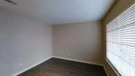 This single-story space presents a straightforward and functional interior designed with simplicity in mind. The room showcased features dark hardwood-style flooring that contrasts cleanly against the white baseboards running along the perimeter, adding a polished, finished look to the space. The walls are painted in a warm, neutral beige tone, creating a versatile backdrop that complements a wide range of furnishing styles and décor choices. The white ceiling is smooth and bright, contributing to an open and airy atmosphere within the room. A single electrical outlet is visible along the lower portion of the wall, indicating practical utility placement throughout. The overall ambiance is calm and understated, with a clean, move-in ready condition that allows occupants to personalize the space freely. The neutral color palette and durable flooring make this space well-suited for a variety of uses, from a living area to a bedroom or home office. The minimalist design ensures that the focus remains on functionality and adaptability.