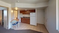 This single-floor unit is designed for efficient, functional living, with an open layout that connects the kitchen and dining areas seamlessly. The kitchen is fitted with warm medium-toned wood cabinetry offering ample storage, complemented by dark countertops and a full suite of white appliances, including a refrigerator, electric range, and an under-cabinet range hood. A double-basin sink is centrally positioned beneath the cabinetry, adding practicality to the workspace. The flooring throughout the kitchen and dining area consists of light beige ceramic tile, providing a clean and easy-to-maintain surface. A small dining area adjacent to the kitchen accommodates a table and chairs, making it a suitable space for everyday meals. The unit transitions from tile to carpet toward the entryway, where a door provides access to the exterior. A doorway visible from the main living area leads to a bathroom equipped with a bathtub, adding convenience to the layout. The overall ambiance is practical and straightforward, with flush-mount ceiling lighting providing adequate illumination throughout the space.