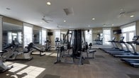 Located at 4610 Weatherford Ln in Columbus, Ohio, this single-floor fitness center is thoughtfully designed to support a comprehensive workout experience. The space is equipped with a wide array of exercise machines, including a multi-station cable weight system, elliptical trainers, treadmills, and a flat utility bench, catering to both strength training and cardiovascular exercise. Large windows line the walls, flooding the room with natural light and offering views of the surrounding outdoor landscape. Full-length mirrors are strategically placed throughout the space, enhancing the sense of openness while allowing users to monitor their form during workouts. The flooring consists of durable, low-pile carpet with defined workout zones, providing both comfort and functionality underfoot. Ceiling fans are installed throughout, promoting air circulation and a comfortable environment during exercise. Recessed lighting supplements the natural light, ensuring the space is well-lit at all times. The neutral color palette of warm beige tones and dark equipment creates a clean, professional ambiance well-suited for focused training sessions.