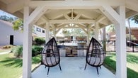 Located in Pensacola, this single-story outdoor amenity space is designed for relaxed communal living and al fresco entertaining. The centerpiece of the space is a generously sized covered pavilion, constructed with striking white-painted timber beams and an exposed truss ceiling that adds architectural character and a sense of openness. Beneath the pavilion, a built-in outdoor kitchen features a stone-clad base equipped with a stainless steel grill and ample prep space, making it ideal for cookouts and gatherings. Two statement teardrop-style wicker lounge chairs flank the cooking area, providing comfortable seating with a stylish, resort-inspired aesthetic. The concrete patio extends seamlessly under the covered structure, offering a clean and durable surface throughout. Just beyond the pavilion, a community pool and sun deck are visible, framed by a wrought-iron fence and lush landscaping including mature palm trees that evoke a distinctly Florida ambiance. Surrounding greenery and the presence of neighboring buildings suggest this space is part of a larger residential community. Natural light floods the area, and the open-air design encourages a connection with the outdoors while still providing ample shade and shelter.