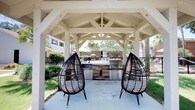 Located in Pensacola, this single-story outdoor amenity space is designed for relaxed communal living and al fresco entertaining. The centerpiece of the space is a generously sized covered pavilion, constructed with striking white-painted timber beams and an exposed truss ceiling that adds architectural character and a sense of openness. Beneath the pavilion, a built-in outdoor kitchen features a stone-clad base equipped with a stainless steel grill and ample prep space, making it ideal for cookouts and gatherings. Two statement teardrop-style wicker lounge chairs flank the cooking area, providing comfortable seating with a stylish, resort-inspired aesthetic. The concrete patio extends seamlessly under the covered structure, offering a clean and durable surface throughout. Just beyond the pavilion, a community pool and sun deck are visible, framed by a wrought-iron fence and lush landscaping including mature palm trees that evoke a distinctly Florida ambiance. Surrounding greenery and the presence of neighboring buildings suggest this space is part of a larger residential community. Natural light floods the area, and the open-air design encourages a connection with the outdoors while still providing ample shade and shelter.