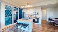 This thoughtfully designed 2-bedroom, 2-bathroom residence offers a contemporary open-plan layout spread across a single floor. The combined kitchen and living area serves as the heart of the unit, where expansive floor-to-ceiling windows flood the space with natural light and frame striking panoramic views of the surrounding urban landscape. The modern kitchen is outfitted with white cabinetry, a subway tile backsplash, stainless steel appliances including a full-size refrigerator, range, microwave, and dishwasher, along with a central island with bar seating and a distinctive curved pendant light fixture. The living area extends seamlessly from the kitchen, offering generous space for seating and direct access to the sweeping city views. The primary bedroom features large corner windows that maximize natural light and provide impressive outdoor sightlines. The second bedroom is equally well-appointed, with access to natural light and views of the exterior. Both bathrooms are finished in a clean, minimalist style with white cabinetry, quartz countertops, and modern fixtures; one includes a glass-enclosed walk-in shower with white tile surround. Wood-look flooring runs throughout the living areas and hallway, while light tile finishes the bathrooms, creating a cohesive and contemporary aesthetic throughout.
