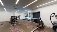 This fitness center offers a comprehensive workout environment designed to support a wide range of exercise routines. The space is laid out across a single floor and features a distinctive geometric-patterned carpet in warm earth tones that adds visual character throughout. Crisp white walls are complemented by bold navy blue accent walls, creating a modern and energizing atmosphere.

The cardio zone is equipped with multiple treadmills positioned near large windows, allowing natural light to fill the area during use. The strength training section houses several Life Fitness cable and weight machines, along with a multi-function rack and adjustable benches, providing options for both targeted and compound exercises. A dedicated free weights area includes a tiered rack stocked with kettlebells and medicine balls, ideal for functional and dynamic training.

A built-in niche along one wall serves as an amenity station, featuring organized storage for yoga and exercise mats, a small beverage refrigerator, and cabinet storage — catering to both flexibility training and hydration needs. Linear LED lighting runs across the ceiling in clean, parallel lines, ensuring bright and even illumination throughout the space. Access to the fitness center is through glass-paneled doors, maintaining an open and connected feel with the surrounding corridor.