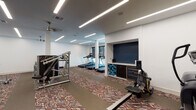 This fitness center offers a comprehensive workout environment designed to support a wide range of exercise routines. The space is laid out across a single floor and features a distinctive geometric-patterned carpet in warm earth tones that adds visual character throughout. Crisp white walls are complemented by bold navy blue accent walls, creating a modern and energizing atmosphere.

The cardio zone is equipped with multiple treadmills positioned near large windows, allowing natural light to fill the area during use. The strength training section houses several Life Fitness cable and weight machines, along with a multi-function rack and adjustable benches, providing options for both targeted and compound exercises. A dedicated free weights area includes a tiered rack stocked with kettlebells and medicine balls, ideal for functional and dynamic training.

A built-in niche along one wall serves as an amenity station, featuring organized storage for yoga and exercise mats, a small beverage refrigerator, and cabinet storage — catering to both flexibility training and hydration needs. Linear LED lighting runs across the ceiling in clean, parallel lines, ensuring bright and even illumination throughout the space. Access to the fitness center is through glass-paneled doors, maintaining an open and connected feel with the surrounding corridor.