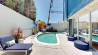Located at 2727 Edison St in San Mateo, California, this thoughtfully designed outdoor amenity space delivers a resort-inspired atmosphere in the heart of the Bay Area. The centerpiece is a sparkling, generously sized swimming pool with crystal-clear blue water, framed by a wide, sun-drenched concrete deck outfitted with rows of navy blue chaise lounges — ideal for relaxing under the California sun.

Floor-to-ceiling glass walls along one side of the pool area create a seamless indoor-outdoor connection, allowing natural light to flood into the adjacent interior lounge space while offering poolside views from within. Bistro-height seating and accent furniture in teal and warm tones add a playful, contemporary touch to the deck.

A vibrant, large-scale California-themed mural anchors one end of the pool courtyard, depicting palm trees, colorful succulents, and a warm sunset palette — giving the space a distinct, energetic identity. Lush planters with tropical and desert plants are arranged throughout to enhance the outdoor ambiance.

A separate, more intimate lounge nook features a linear fire table, woven outdoor furniture, and decorative lattice privacy screens with geometric patterns, offering a quieter retreat. The multi-story building facade, clad in bold blue and warm wood tones, completes the architecturally cohesive outdoor environment.