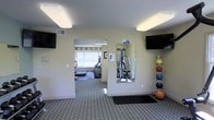 This fitness center is designed to provide a comprehensive workout experience within a single-floor layout. The space is divided into two connected areas, each serving a distinct purpose for exercise and training. The main area features durable carpeted flooring, wall-mounted flat-screen televisions for entertainment during workouts, and a large mirror that enhances the sense of depth while allowing users to monitor their form. A medicine ball rack stocked with a colorful assortment of weighted balls adds a functional free-weight element to the space. An adjoining cardio room, visible through an open passage, is bathed in natural light from a large window and houses a selection of cardio machines, including an elliptical and a treadmill. A multi-function cable and strength training machine is also present, offering a variety of resistance exercises. The walls are painted in a neutral, light tone that contributes to a clean and open ambiance. Flush ceiling lights provide consistent, even illumination throughout both areas. The overall layout is thoughtfully organized to accommodate a range of fitness activities, from cardiovascular training to strength and functional movement, making it a versatile and practical workout facility.