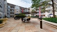 Located at 500 Bedford St in Stamford, Connecticut, this courtyard space offers a welcoming outdoor environment nestled within a multi-story residential complex. The expansive patio is finished with large-format stone-look tiles and is furnished with multiple dining sets, each shaded by generous market umbrellas, creating comfortable gathering areas for residents and guests. Mature trees and well-maintained landscaping line the perimeter, providing natural shade and a sense of privacy. Decorative bollard lighting is positioned throughout the courtyard, contributing to both safety and ambiance. The surrounding building features balconied residential units that overlook the courtyard, reinforcing a sense of community.

The space also includes a public restroom finished with a durable combination of beige ceramic tile wainscoting and soft blue upper walls. The flooring consists of neutral ceramic tile throughout. The restroom is fully ADA-compliant, equipped with stainless steel grab bars near the toilet, a wall-mounted accessible sink, a paper towel dispenser, soap dispenser, and a paper seat cover unit. A decorative potted plant adds a touch of warmth to the space. The restroom is designed to be both functional and accessible, serving the needs of all courtyard visitors.