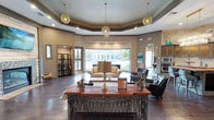 This refined single-floor space makes a striking first impression with its grand, open layout and thoughtfully curated design elements. The central lounge area is anchored by a dramatic tray ceiling adorned with decorative molding, from which hang three ornate gold globe pendant lights that cast a warm, inviting glow throughout the room. Dark hardwood-style flooring runs throughout, providing a rich contrast to the lighter wall tones and furnishings.

A decorative wrought iron console table with a rich wood top serves as a focal centerpiece, accented by potted greenery and tasteful décor. Seating areas are arranged to encourage both comfort and conversation, featuring plush upholstered sofas, tufted leather chairs, and coordinating accent pieces. Large floor-to-ceiling windows and glass doors at the rear of the space flood the interior with natural light while offering sweeping views of lush outdoor landscaping.

Additional areas visible from the main lounge include a secondary lounge or game room with a pool table, as well as what appears to be a kitchen or bar area equipped with built-in appliances. Arched doorways and varied architectural details contribute to the space's polished, upscale ambiance, making it a welcoming and visually impressive environment throughout.