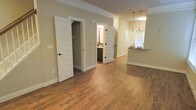 This two-story space showcases a thoughtfully designed interior that balances modern aesthetics with functional living. The main level features warm-toned hardwood-style flooring that extends throughout the open layout, creating a cohesive and inviting atmosphere. Neutral beige walls complement the light-colored trim and white five-panel doors with dark hardware accents, adding a subtle contrast that enhances the overall design.

The open floor plan connects the main living area to the kitchen, which is visible in the background and distinguished by pendant lighting that hangs above what appears to be a countertop bar or island. The kitchen benefits from natural light streaming through a window, brightening the space. Crown molding along the ceiling adds an architectural detail that elevates the interior's finish quality.

Two doorways visible from the main living area lead to additional rooms, including what appears to be a bathroom with dark cabinetry and a sink. The layout suggests a well-organized flow between spaces. With its clean lines, consistent material choices, and attention to detail, this two-floor property presents a polished, move-in-ready environment suited for comfortable everyday living.