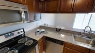 This single-floor space showcases a thoughtfully designed kitchen that serves as a central highlight. The kitchen features rich, dark-toned wooden cabinetry that extends to the upper walls, providing generous storage throughout. Granite-style countertops in a neutral speckled finish offer ample workspace and lend a clean, polished aesthetic to the area. The kitchen is equipped with a full suite of appliances, including a black electric coil range, a stainless steel over-the-range microwave, a built-in dishwasher, and a sink positioned along the perimeter. The cabinetry hardware features sleek, brushed metal pulls that complement the stainless steel appliances cohesively. The layout is designed in an efficient L-shape, maximizing usable counter space while maintaining an open flow. Light-toned flooring provides a subtle contrast to the darker cabinetry, brightening the overall atmosphere. Natural light enters through a nearby window, adding warmth to the space. The overall ambiance is functional yet refined, with a consistent design palette that creates a well-coordinated and practical kitchen environment suitable for everyday use.