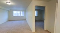 This single-floor residential space offers a clean, functional layout with a calm and neutral aesthetic throughout. The main living area is generously proportioned, featuring plush wall-to-wall carpeting in a warm beige tone that adds comfort underfoot. Soft, neutral-painted walls create a bright and airy atmosphere, complemented by natural light streaming in through large windows that frame views of the surrounding exterior landscape. The open feel of the main living space is enhanced by its unobstructed layout, making it versatile for a variety of furniture arrangements. A doorway leads to an adjacent room — likely a bedroom — which mirrors the design language of the main area, with matching carpet, neutral walls, and its own window allowing for continued natural light penetration. The consistent finishes across both spaces create a cohesive and understated interior. Baseboard heating elements are visible along the walls, indicating a practical and efficient heating system. The overall ambiance is quiet and move-in ready, offering a blank canvas that lends itself well to personalization. The space is well-suited for those seeking a straightforward, comfortable residential environment with an emphasis on light and simplicity.