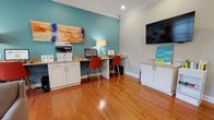 Located at 1341 Ellis Forest Rd in Kernersville, North Carolina, this single-floor amenity facility offers residents two well-appointed shared spaces designed for productivity and wellness.

The business center features warm hardwood flooring, white cabinetry with granite countertops, and a built-in workstation with dual computer monitors and seating for two. A multifunction printer, wall-mounted flat-screen TV, and colorful abstract artwork complete the space, all set against a vibrant teal accent wall and crisp white crown molding that lends a polished, professional atmosphere.

The fitness center is generously equipped for a full range of workouts. Large floor-to-ceiling windows flood the space with natural light and provide views of the surrounding greenery and pool area. Cardio equipment includes elliptical machines and treadmills, while the strength training area offers a multi-station cable machine, a Smith machine, a dumbbell rack with a full weight selection, a weight bench, and exercise stability balls. Full-length mirrors amplify the sense of space, and durable rubber flooring ensures a safe workout environment. French doors provide direct access to the outdoor area. The consistent teal and light-blue color palette, crown molding, and recessed lighting create a cohesive, welcoming atmosphere throughout both areas.