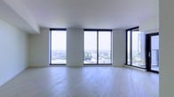 This contemporary space makes an immediate impression with its clean, minimalist design and an abundance of natural light. The most striking feature is the expansive floor-to-ceiling windows framed in bold black trim, which span nearly the entire perimeter of the main living area. These oversized windows offer sweeping panoramic views of the cityscape below, including tree-lined streets and the surrounding urban landscape stretching to the horizon — a breathtaking backdrop at any time of day.

The interior is finished with light-toned hardwood flooring that runs throughout, lending warmth and continuity to the space while complementing the bright, airy atmosphere created by the generous glazing. The walls and ceiling are rendered in a clean white, reinforcing the sense of openness and allowing the views and natural light to take center stage.

The layout is open and unencumbered, offering flexibility for a variety of uses and configurations. The elevated position of the space amplifies the feeling of being immersed in the cityscape, making it feel both expansive and connected to the world outside. Overall, the space exudes a refined, modern sensibility defined by its architectural clarity, luminosity, and commanding outlook.