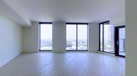 This contemporary space makes an immediate impression with its clean, minimalist design and an abundance of natural light. The most striking feature is the expansive floor-to-ceiling windows framed in bold black trim, which span nearly the entire perimeter of the main living area. These oversized windows offer sweeping panoramic views of the cityscape below, including tree-lined streets and the surrounding urban landscape stretching to the horizon — a breathtaking backdrop at any time of day.

The interior is finished with light-toned hardwood flooring that runs throughout, lending warmth and continuity to the space while complementing the bright, airy atmosphere created by the generous glazing. The walls and ceiling are rendered in a clean white, reinforcing the sense of openness and allowing the views and natural light to take center stage.

The layout is open and unencumbered, offering flexibility for a variety of uses and configurations. The elevated position of the space amplifies the feeling of being immersed in the cityscape, making it feel both expansive and connected to the world outside. Overall, the space exudes a refined, modern sensibility defined by its architectural clarity, luminosity, and commanding outlook.