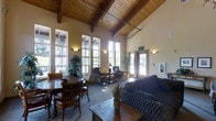 This single-story common lounge showcases striking architectural design highlighted by soaring vaulted ceilings finished with warm wood planking and prominent exposed timber beams. The elevated ceiling height creates an expansive, airy atmosphere throughout the space. Tall, multi-pane windows line the walls, flooding the interior with generous natural light and offering views of the surrounding greenery outside.

The space is thoughtfully organized into two distinct seating areas. On one side, a traditional dining arrangement features a round wooden table paired with classic ladder-back chairs, ideal for casual meals or group gatherings. On the opposite side, a comfortable lounge area is furnished with a plush, patterned sofa and coordinating armchairs, centered around a carved wooden coffee table — perfect for relaxed conversation.

A cozy seating vignette with wicker chairs and a small side table occupies a transitional zone between the two areas. Potted indoor plants are strategically placed throughout, adding organic texture and warmth. Wall sconces provide soft ambient lighting to complement the natural light. The neutral, earthy color palette and combination of wood and fabric finishes contribute to an inviting, lodge-like ambiance.