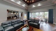 Located at 9611 Grant Rd in Houston, TX, this single-floor community clubhouse is thoughtfully designed to provide a comfortable and functional gathering space. The main lounge area immediately impresses with its elevated tray ceilings accented by a dark finish and a decorative chandelier, creating a refined yet inviting atmosphere. Rich hardwood-style flooring runs throughout, complementing the neutral gray walls and contemporary furnishings, including plush leather sofas and accent chairs arranged around a wooden coffee table. Colorful abstract artwork adorns the walls, adding visual interest throughout the space. A beverage station with a dark wood credenza provides convenient amenity access for guests. The dining area is a standout feature, positioned beside floor-to-ceiling windows and a glass sliding door that frame lush, landscaped outdoor views with tropical palms, flooding the space with natural light. Teal drapery panels add a pop of color and elegance. The kitchen is well-appointed with white cabinetry, dark granite countertops, stainless steel appliances including a refrigerator and dishwasher, and a center island — ideal for community events and gatherings. The arched entryway and open flow between zones make this clubhouse both architecturally appealing and highly practical.